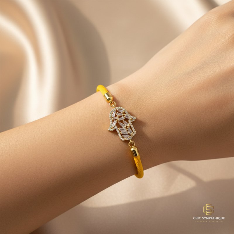 Poignet d'une femme portant un bracelet en cordon jaune vif. Le pendentif est une main de Fatima (Hamsa) dorée, avec un contour pavé de petits cristaux et un centre serti de cristaux blancs taille baguette.
