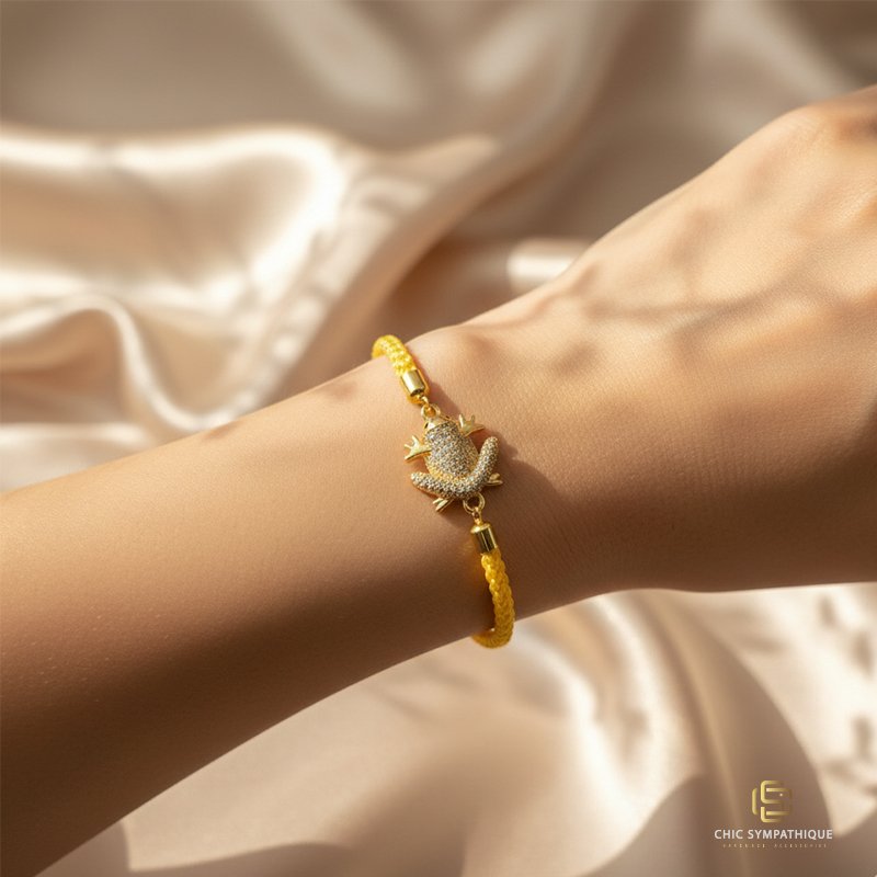 Poignet d'une femme portant un bracelet en cordon tressé jaune. Le pendentif est une grenouille stylisée, dorée et entièrement pavée de cristaux blancs scintillants.