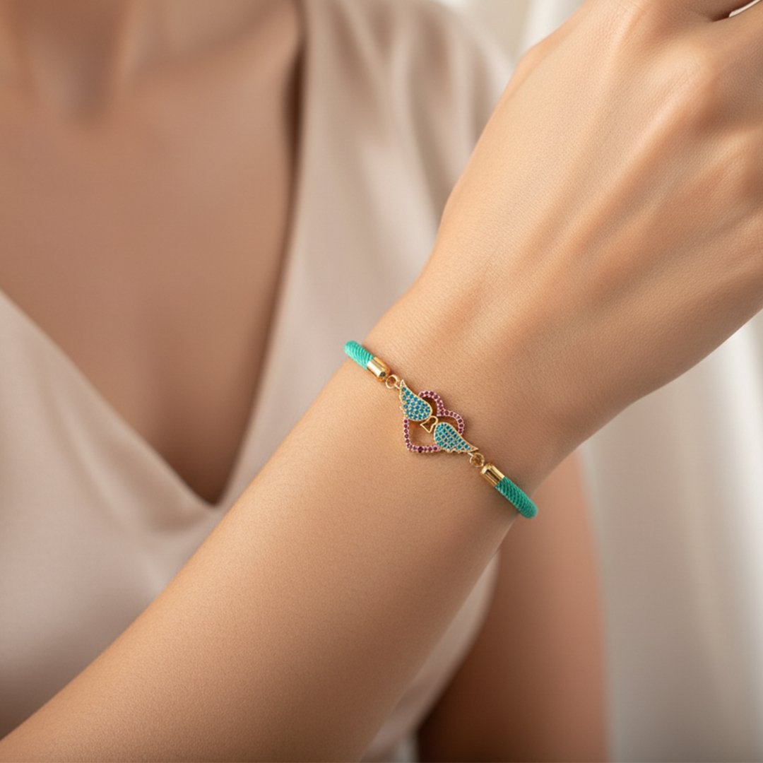 Poignet d'une femme portant un bracelet en cordon turquoise. Le pendentif est une pièce dorée en forme de deux cœurs entrelacés, l'un pavé de cristaux turquoise et l'autre de cristaux fuchsia, formant une silhouette d'ailes.